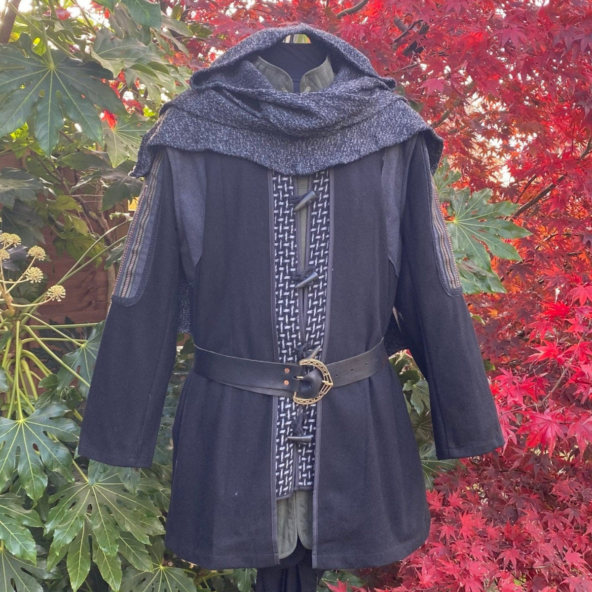 Medieval LARP tunic with patterned trim and buckle belt, leafy backdrop.