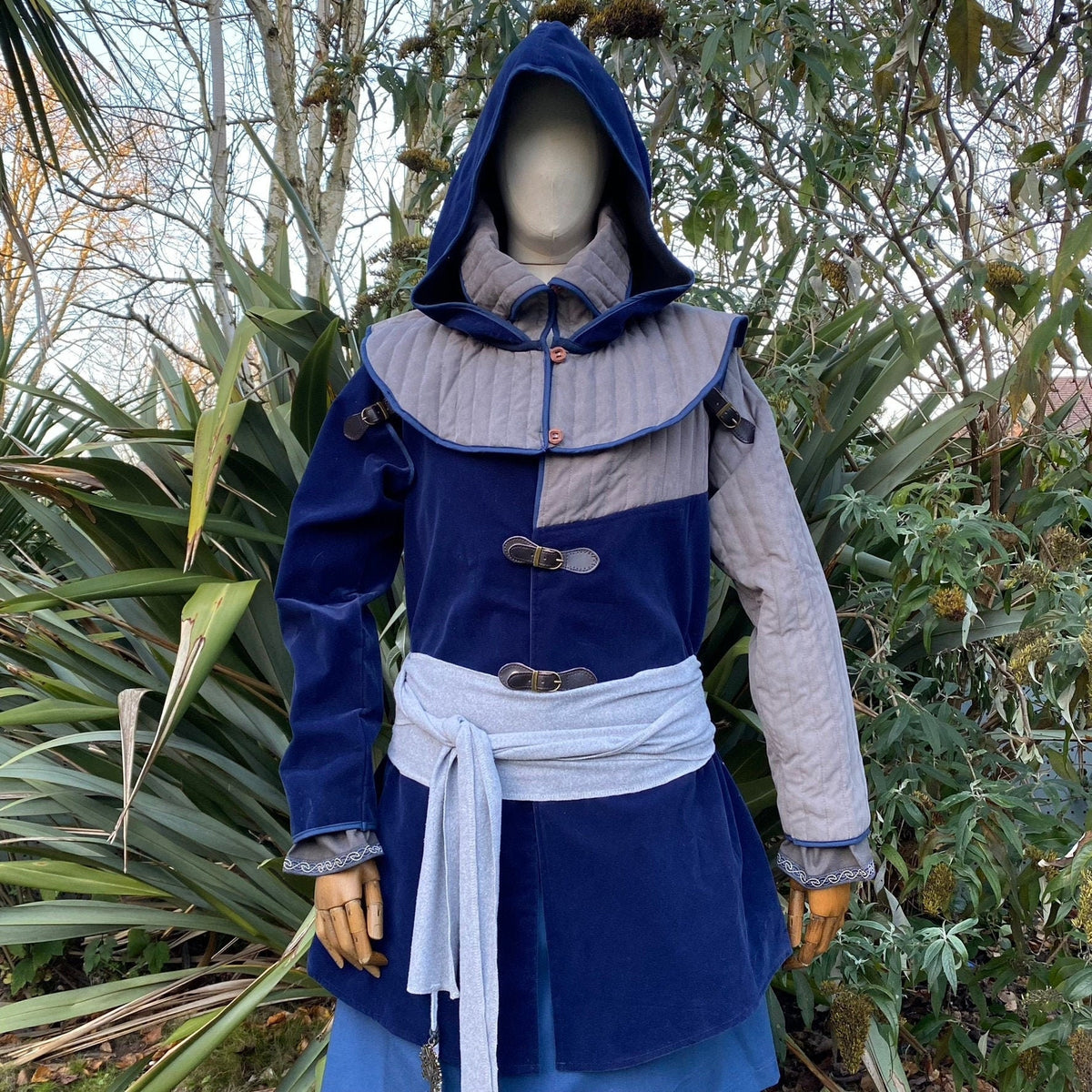Azure medieval LARP costume with hood and belts in a wooded setting.