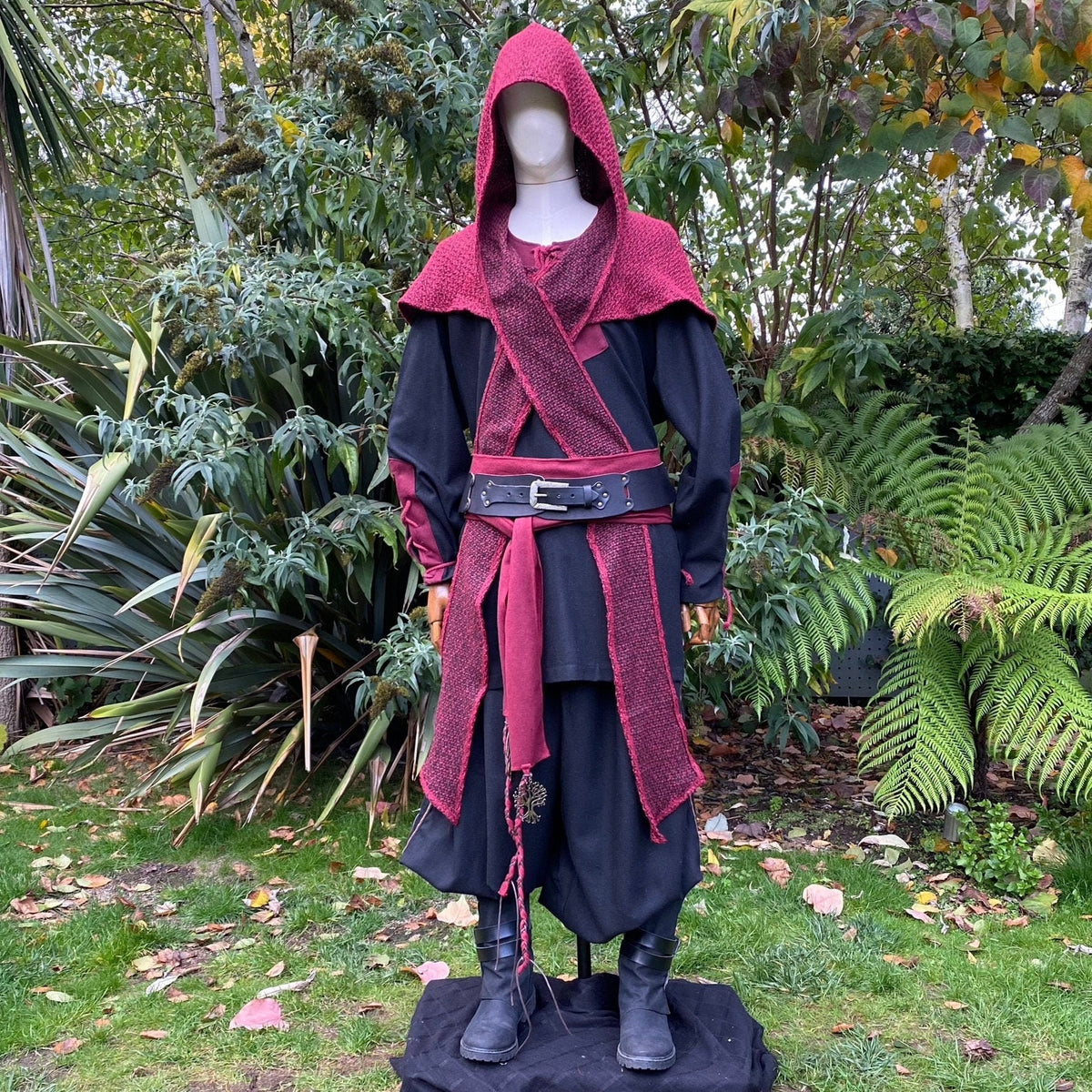 Medieval LARP outfit on mannequin, red hood and sash, lush greenery.